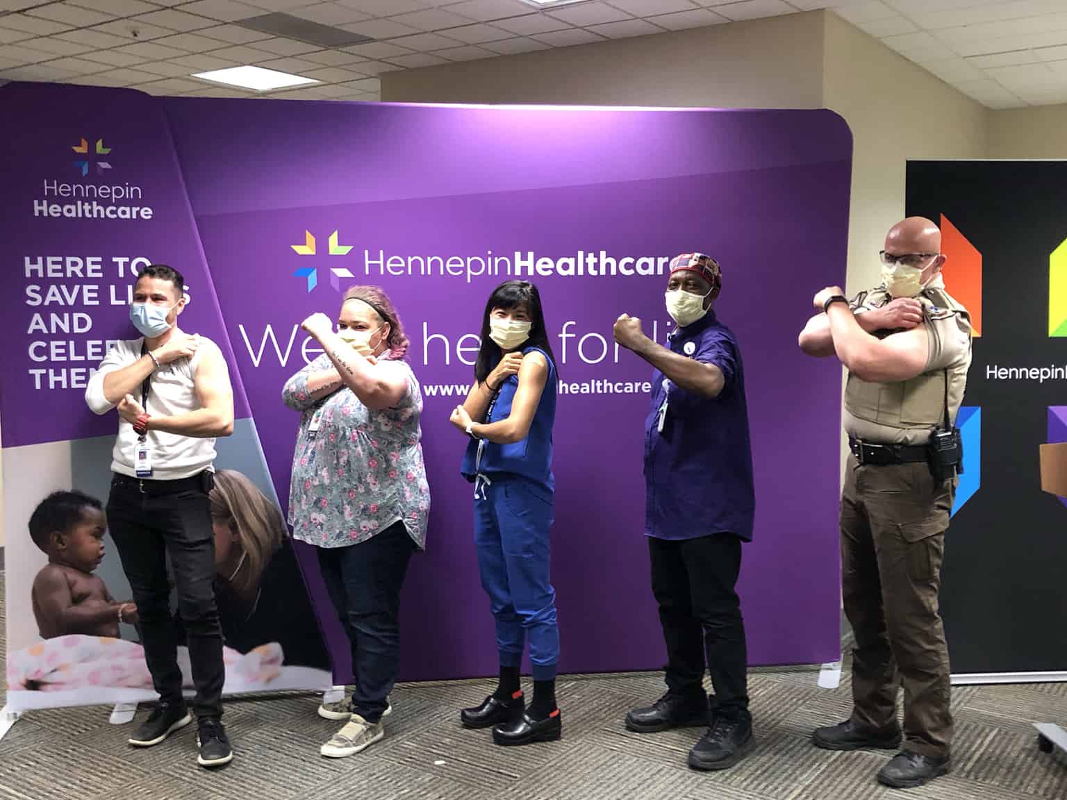 five staff members in front of branded wall holding up arms where they had a vaccine