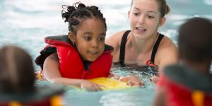 young girl with life preserver with instructor and other children learning to swim, children and water safety, drowning leading cause of death in children, recognition of risk associated with water, floatation device, child water safety