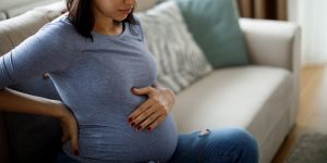 pregnant woman sitting on couch and holding her belly, Women’s health physical therapy is restoration and strength, incontinence, sciatica, pelvic care after pregnancy, labor and delivery position, beth stegora, breastfeeding support