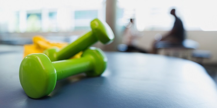 Close up of green hand weights and a yellow resistance band on a table in a rehabilitation clinic. A physical therapist meets with a client in the background.