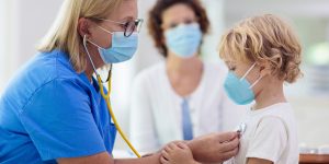 masked doctor listens to boy's heart with stethoscope with nurse in background, pediatric diseases, vaccinating children, common childhood illnesses, flu vaccine, pediatric illnesses